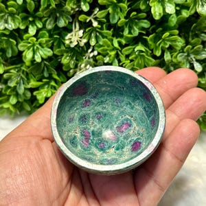May include: A small, round bowl made of green stone with red and pink inclusions. The bowl has a smooth, polished interior and a slightly rougher exterior. The bowl is held in a hand, with a green leafy background.