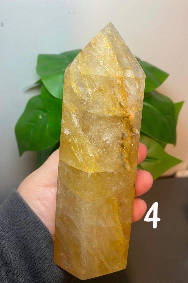 Puede incluir: Una gran punta de cristal de cuarzo transparente con un tono amarillo dorado. El cristal tiene una forma natural facetada y mide aproximadamente 15 cm de alto. El cristal est&aacute; sentado en una mesa con una planta verde en el fondo.