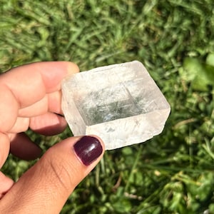 May include: A clear, cube-shaped crystal with visible internal structures. The crystal is held against a backdrop of green grass, showcasing its translucent quality. The edges of the crystal are rough, giving it a natural appearance.