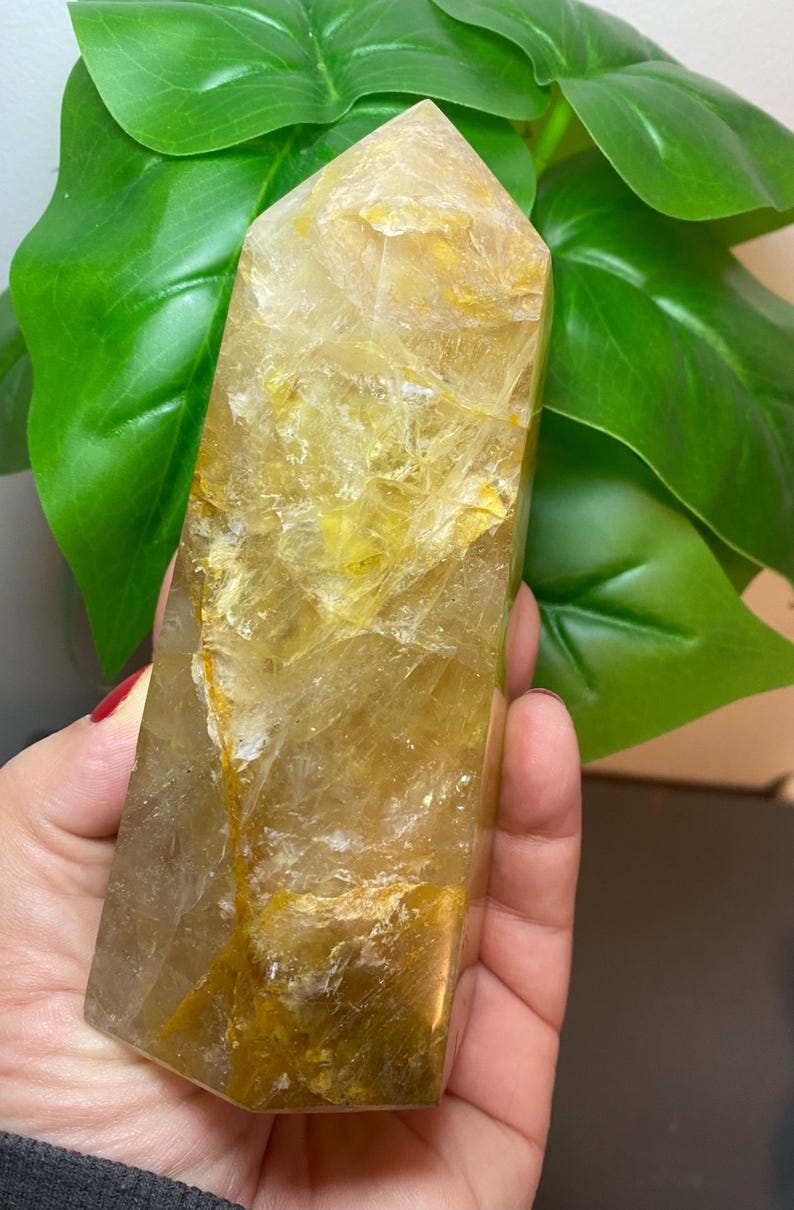 Puede incluir: Una gran punta de cristal de cuarzo transparente con un tono amarillo dorado. El cristal tiene un aspecto natural y bruto con muchas inclusiones y estr&iacute;as.