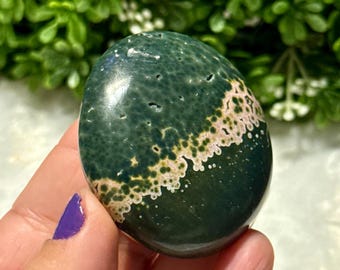May include: A polished, oval gemstone with a dark green base and a diagonal band of beige and white spots. The stone has a smooth, glossy surface. The background is blurred green foliage. The stone is held in a hand with a purple painted fingernail.