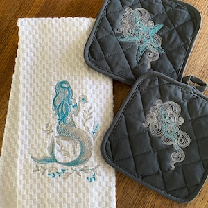 May include: White waffle-weave kitchen towel and two gray quilted pot holders. The towel features an embroidered mermaid in teal and gray. The pot holders have embroidered starfish and sea-themed designs in teal and gray.
