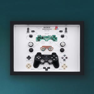 Op de afbeelding: Een ingelijste display met een gedemonteerde Sony PlayStation 2 DualShock 2 controller. De componenten zijn gerangschikt op een witte achtergrond, inclusief de printplaat, knoppen en joystick. De lijst is zwart.
