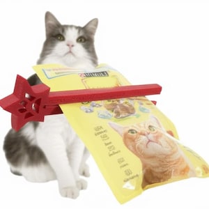 Puede incluir: Un gato gris y blanco con un clip de bolsa rojo en forma de estrella que sostiene una bolsa amarilla de comida para gatos. La bolsa presenta una ilustración de un gato naranja. El gato está mirando hacia arriba.