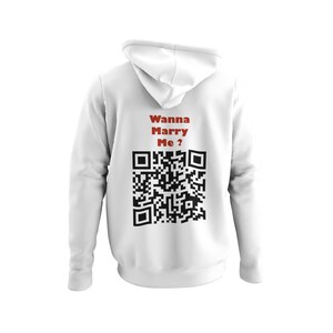 Op de afbeelding: Witte hoodie met de tekst "Wanna Marry Me?" in het rood boven een zwarte QR-code. De hoodie is effen wit van kleur en heeft lange mouwen.