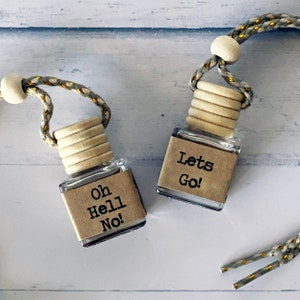 May include: Two small glass bottles with wooden caps and braided cords. One bottle reads "Oh Hell No!" and the other says "Lets Go!" These are likely air fresheners or decorative items.