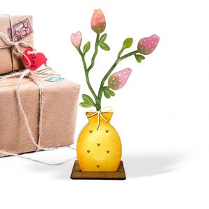 May include: A wooden floral decoration featuring a yellow vase with heart accents, holding a green stem with pink and white dotted flower buds. The vase sits on a brown base. Two wrapped packages are in the background.