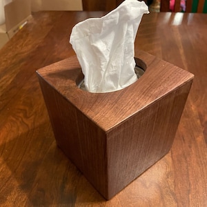 May include: A dark brown wooden tissue box holder with a white tissue box inside. The tissue box holder is on a wooden table.