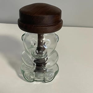 May include: A clear glass bear-shaped container with a dark brown wooden lid. The container has a detailed bear design, including facial features and paws. The lid is a flat, circular shape.