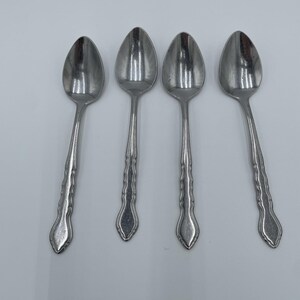May include: Four stainless steel spoons with oval bowls and decorative handles. The spoons are arranged on a white surface, showcasing their polished, reflective surfaces. The handles have a textured, ridged design.