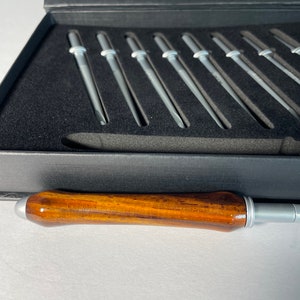 May include: A set of silver knitting needles in a black box with a wooden handle. The needles are arranged in a row and are all the same size.