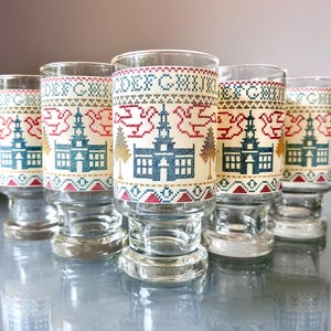 May include: A set of five clear glass tumblers featuring a cross-stitch style pattern. The design includes a repeating alphabet from A to K, stylized church buildings in blue, red birds, and gold trees, all set against a cream-colored band.
