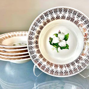 May include: A stack of white ceramic soup bowls featuring a central illustration of a white trillium flower with green leaves. The rim of each bowl is decorated with a repeating brown and gold geometric pattern of stylized fan-like shapes.