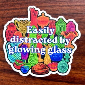 May include: A sticker featuring a collection of colorful glass objects, including vases, bowls, and decorative pieces. The text "Easily distracted by glowing glass" is in the center. The sticker has a white border and is set against a dark wood background.
