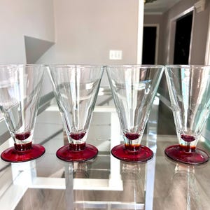 May include: Four clear, cone-shaped glasses with a red base. The glasses are made of clear glass and have a faceted design. The red base is a solid, circular shape. The glasses are arranged in a row on a reflective surface.
