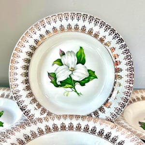 May include: A white ceramic bowl featuring a central illustration of a white trillium flower with green leaves. The rim is decorated with a repeating metallic gold and black filigree pattern. The bowl has a wide, shallow basin and a smooth finish.
