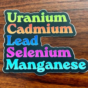 May include: A black sticker with the words "Uranium, Cadmium, Lead, Selenium, Manganese" in colorful, bubble-style letters. The sticker has a white border and is set against a wooden background.