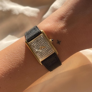 May include: A gold rectangular wristwatch with a black leather strap. The watch face is adorned with numerous small, sparkling stones. This classic watch design is suitable for daily wear or special events.