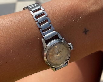 May include: A vintage silver wristwatch with a light-colored face and silver band is shown on a wrist. The watch has a round face with numbers and hands. The band is made of rectangular links. A small tattoo is visible on the wrist.