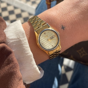 May include: A gold wristwatch with a rectangular face and a gold link strap. The watch face is cream-coloured with gold detailing and shows the day of the week. The watch is worn on a wrist with a small tattoo.