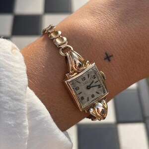 Könnte beinhalten: Eine Vintage-Armbanduhr aus Gold mit quadratischem Zifferblatt und strukturiertem Armband. Das Zifferblatt zeigt schwarze Ziffern auf hellem Hintergrund. Die Uhr wird an einem Handgelenk mit einem kleinen Tattoo getragen.