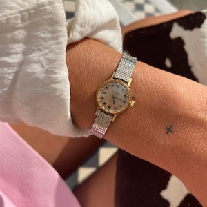 May include: A vintage-style wristwatch featuring a round gold-tone case and a textured gold dial with dark hour markers. The watch is attached to a flexible silver-tone metal mesh strap with a fine, woven pattern that wraps around the wrist.