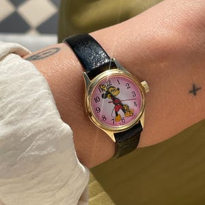 Pode incluir: Um relógio de pulso dourado com uma pulseira de couro preta. O mostrador rosa do relógio exibe algarismos pretos e uma imagem de desenho animado do Mickey Mouse. O relógio está preso ao pulso.