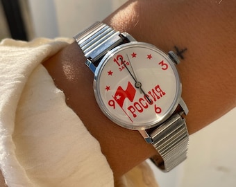 May include: A silver wristwatch with a white face and a red star and flag design. The watch face says "Заря" and "Россия" in Cyrillic lettering.