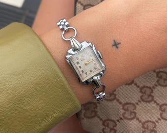 May include: A vintage silver wristwatch with a rectangular face and a light-colored dial. The watch has a delicate, ornate design and is attached to a silver link bracelet. The watch is worn on a wrist with a small tattoo.