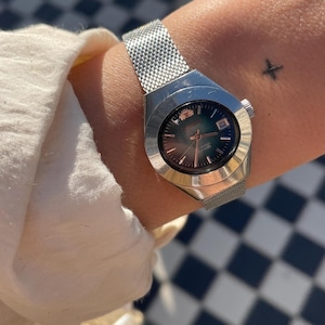 May include: A silver-tone wristwatch featuring a round, dark-colored dial with metallic hour markers and a date window at the three o'clock position. The watch has a polished metal case and a flexible, fine-mesh stainless steel strap.