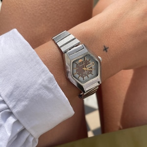 May include: A silver-toned wristwatch with a geometric face and a metal band. The watch has a brown face with gold accents and a day-date display. The watch is worn on a wrist with a small tattoo.