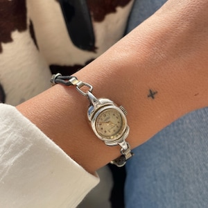May include: A vintage silver wristwatch with a round face and a black leather strap. The watch face has gold numerals and hands. The watch is worn on a wrist with a small tattoo. The background includes a white shirt and blue jeans.