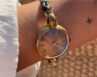 May include: Vintage gold-toned wristwatch with a worn, aged appearance. The watch features a round face with visible numerals and hands, attached to a black leather strap. The watch is worn on a wrist.
