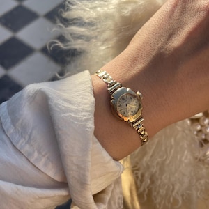 Op de afbeelding: Een goudkleurig polshorloge met een bloemvormige wijzerplaat en een schakelband. De wijzerplaat van het horloge toont cijfers en wijzers. Het horloge wordt om de pols gedragen, met een witte mouw en een pluizig, crèmekleurig kledingstuk op de achtergrond.