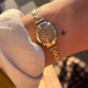 May include: A vintage gold-toned Premier wristwatch with a floral-shaped face and a linked band. The watch face displays the Premier brand name and the words "17 Jewels". The watch is worn on a wrist.