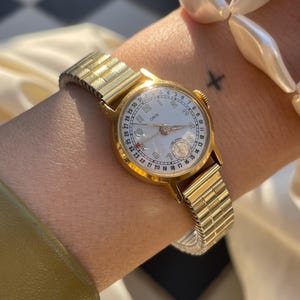 Może przedstawiać: Złoty zegarek na rękę z białą tarczą i czarnymi cyframi. Zegarek ma złotą bransoletę, a na tarczy widnieje napis "ORIS". Zegarek jest na nadgarstku.