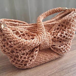 May include: A peach-colored crochet handbag featuring a large, prominent bow design on the front. The bag is constructed with an open-weave lattice pattern and includes a single handle attached with gold-toned metal hardware on both sides.