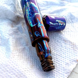 Puede incluir: Una pipa de metal con un diseño de hongo en tonos azules, morados y bronce. La pipa tiene una base roscada y una tapa desmontable, ambas con la misma obra de arte caprichosa de hongos. El acabado iridiscente le da un aspecto único.