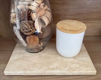 May include: A clear glass vase filled with dried botanicals, including wood curls and seed pods, sits on a beige stone tile. A white ceramic candle with a wooden lid, marked "aromabotanica", is next to the vase.