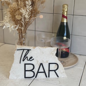 May include: A white stone sign with the words "The BAR" in black lettering. The sign is sitting on a tiled surface with a bottle of champagne and two glasses in the background.
