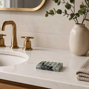 May include: A rectangular soap dish crafted from green marble with a wavy, grooved surface texture. The soap dish features natural dark veining throughout the stone, providing a polished finish that sits on a white countertop surface.