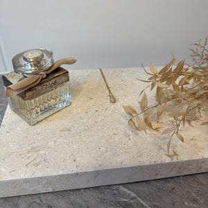 Op de afbeelding: Een gouden ketting met een kleine hanger en een parfumflesje met de merknaam "Chloé" worden getoond op een lichtgekleurde rechthoekige tray. Gedroogde planten zijn aan de rechterkant van de tray geplaatst.
