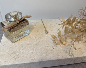 May include: A gold necklace with a small pendant and a perfume bottle with the brand name "Chloé" are displayed on a light-colored rectangular tray. Dried plants are arranged on the right side of the tray.