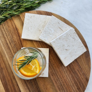 May include: Four white marble coasters with a light brown wood background. A glass with ice, a slice of orange, and rosemary is sitting on one of the coasters.