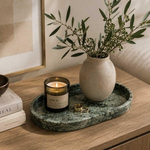 May include: An oval green marble tray holding a lit candle in a dark glass jar with a label reading Santal 26, a beige ceramic vase containing green leafy branches, and two gold-toned rings. The tray is positioned on a light wood surface.