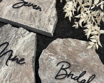 Può includere: Tre segnaposto in pietra grigia con calligrafia nera. I segnaposto dicono "Seven", "Nine" e "Bridal".