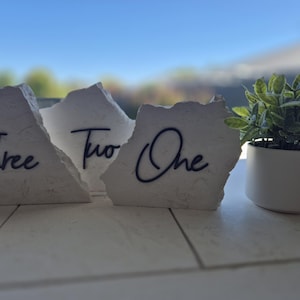 Puede incluir: Tres números de mesa de piedra blanca con textura rugosa y caligrafía negra. Los números son "Uno", "Dos" y "Tres". Una pequeña planta verde en maceta está en el fondo.