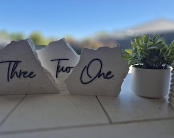 Pode incluir: Três números de mesa de pedra branca com textura áspera e caligrafia preta. Os números são "Um", "Dois" e "Três". Uma pequena planta verde em vaso está no fundo.