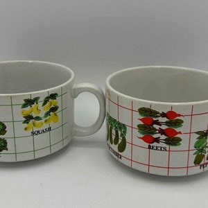May include: Two white ceramic mugs with a grid pattern and colorful illustrations of vegetables. The mugs feature the words "Lettuce", "Squash", "Cucumber", "Beets", and "Peppers".