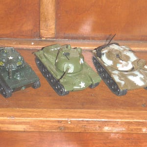 May include: Three toy tanks in olive green and camouflage patterns. The tanks have black treads and white star markings. One tank has the text "U.S. ARMY" on the side. The tanks are on a wooden surface.